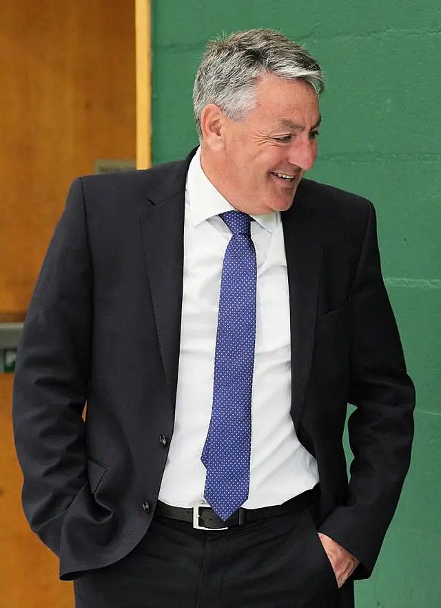 Man in black suit with blue tie is laughing at something out of shot. He has his hands in his pocket and his suit jacket unbuttoned