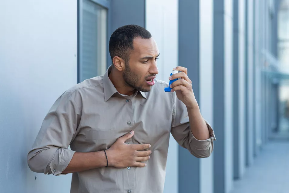 A man outside an office building has a severe asthma attack, a businessman is having difficulty breathing, a worker in a shirt is using an inhaler to make breathing easier, a hispanic man in a casual shirt.