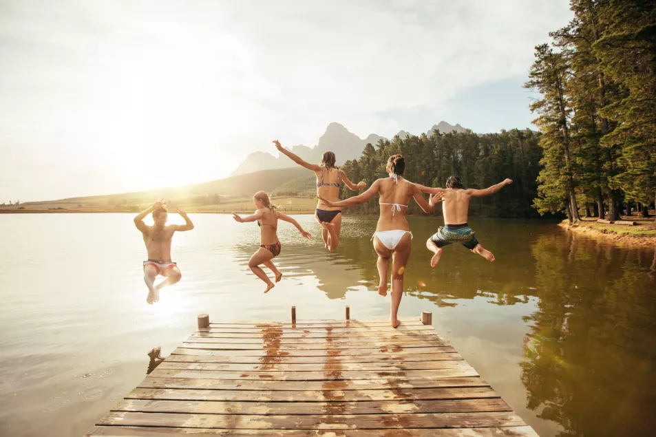 Group of friends jumping into a beautiful lake off a wooden jetty, with mountains and forests on the horizon 