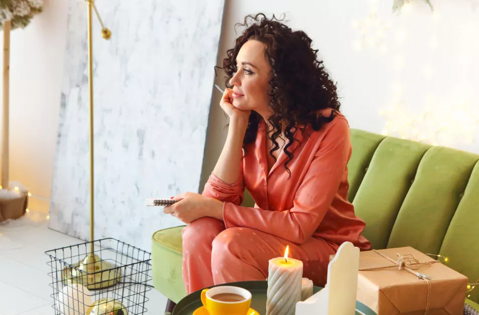 Woman sitting on a green sofa, wearing pyjamas, writing in a notebook. Next to her, a candle, cup of tea and boxed-up gift sit on a coffee table