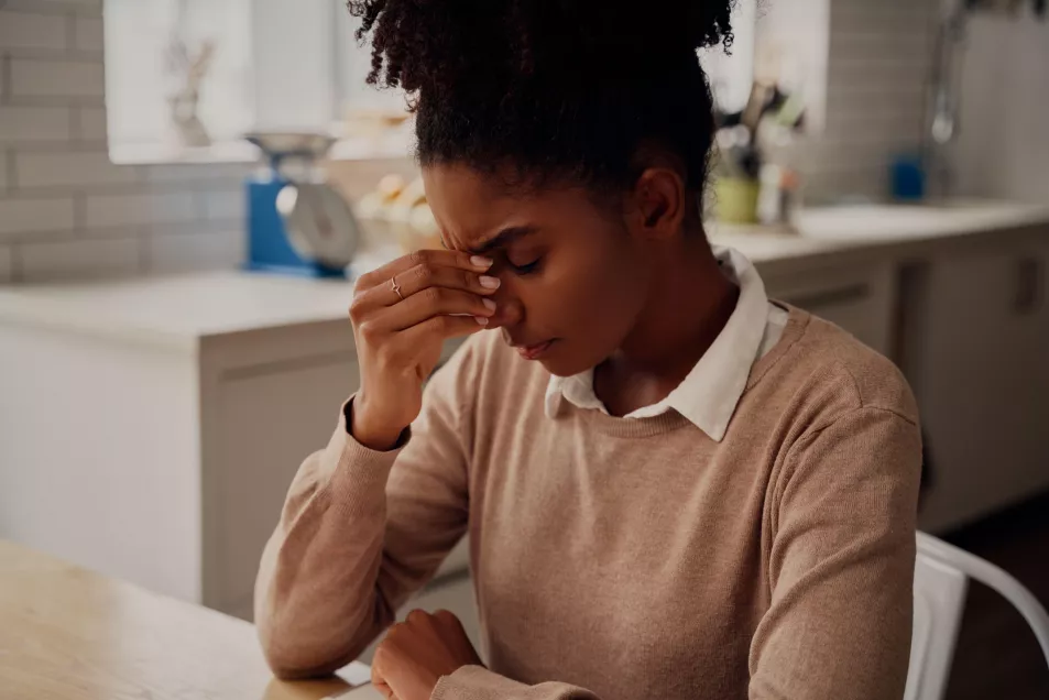 Young black woman sitting in the kitchen at home, touching her forehead and looking very tired