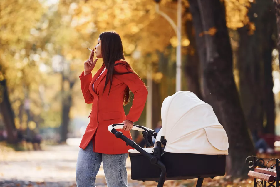 Mother in red coat have a walk with her kid in the pram in the park at autumn time and smoking