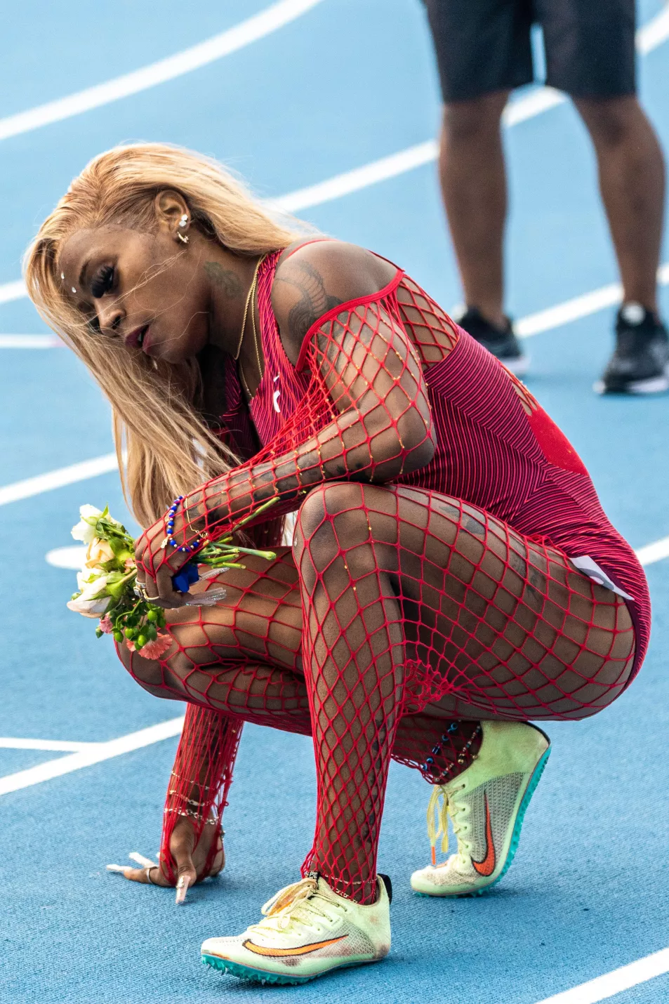 Richardson bends down to pick up flowers in maroon fishnet catsuit and bodysuit on the track after winning a 200 metre sprint