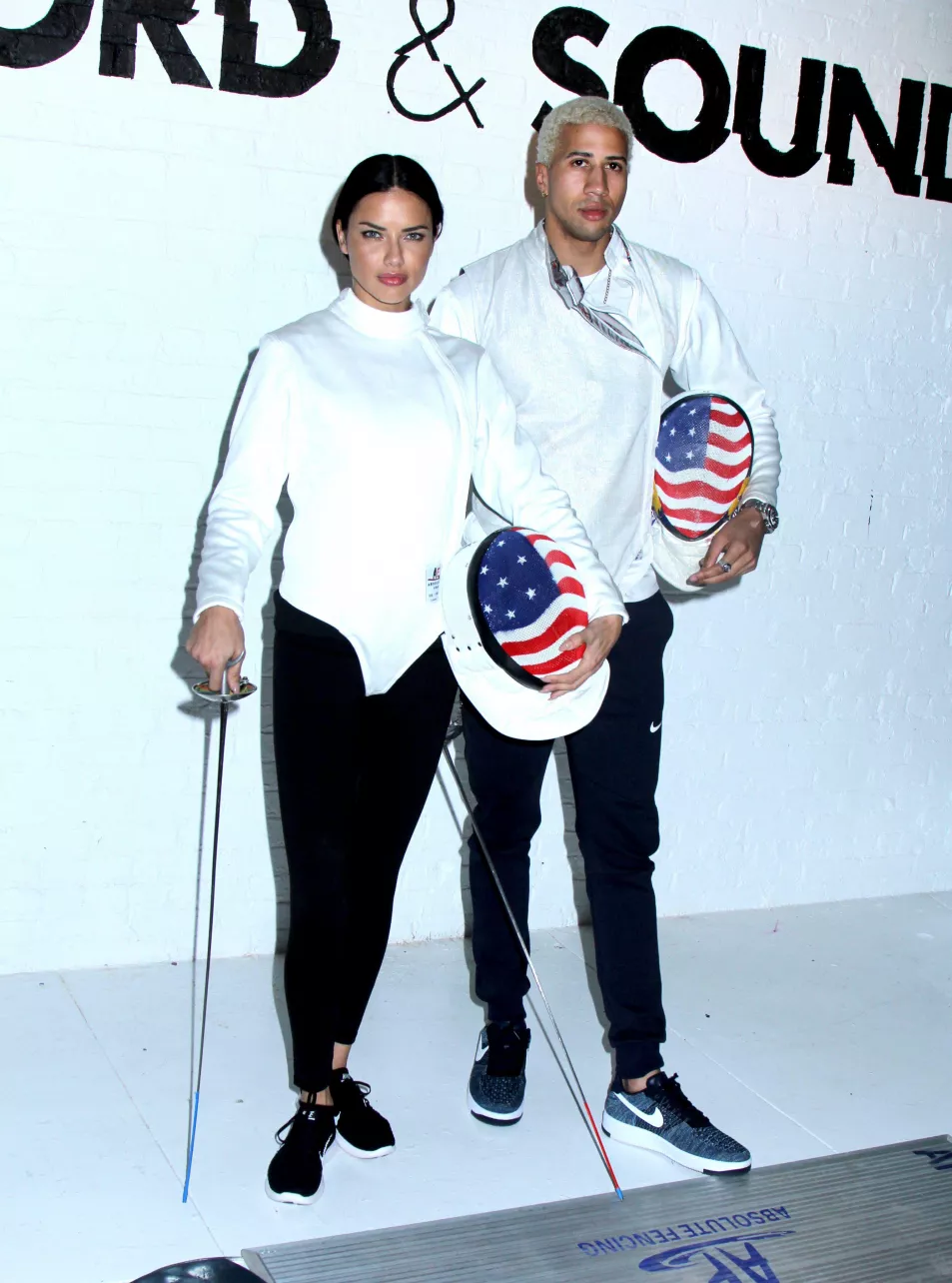 Adriana Lima and Miles Chamley-Watson pose in fencing gear with Amercian flag embellishedhelmets