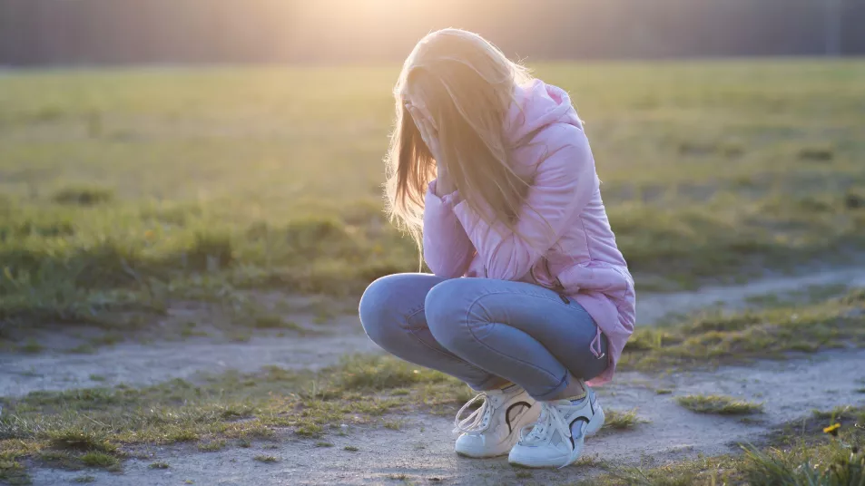 Teenage girl squatting and crying in a field with a sunset in the background