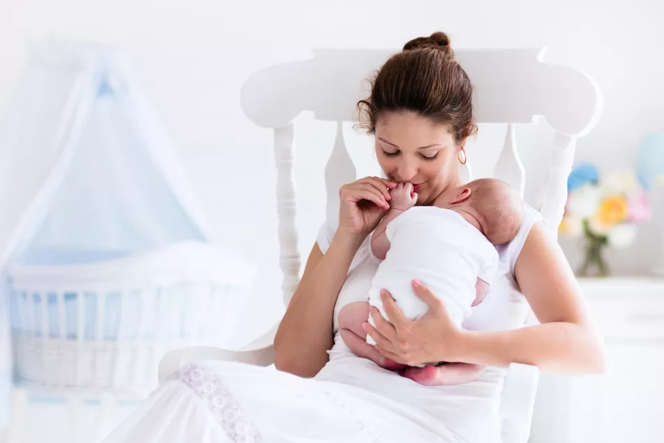 Young mother holding her newborn child. Mom nursing baby. Woman and new born boy relax in a white bedroom with rocking chair