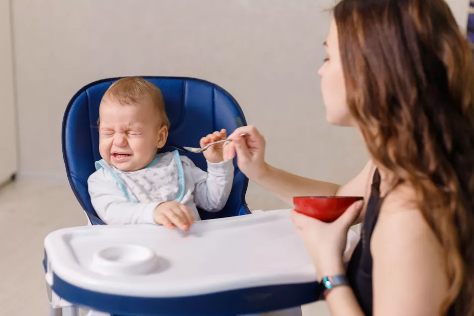 A baby sat in a high chair making disgusted faces at his mum who is trying to feed him