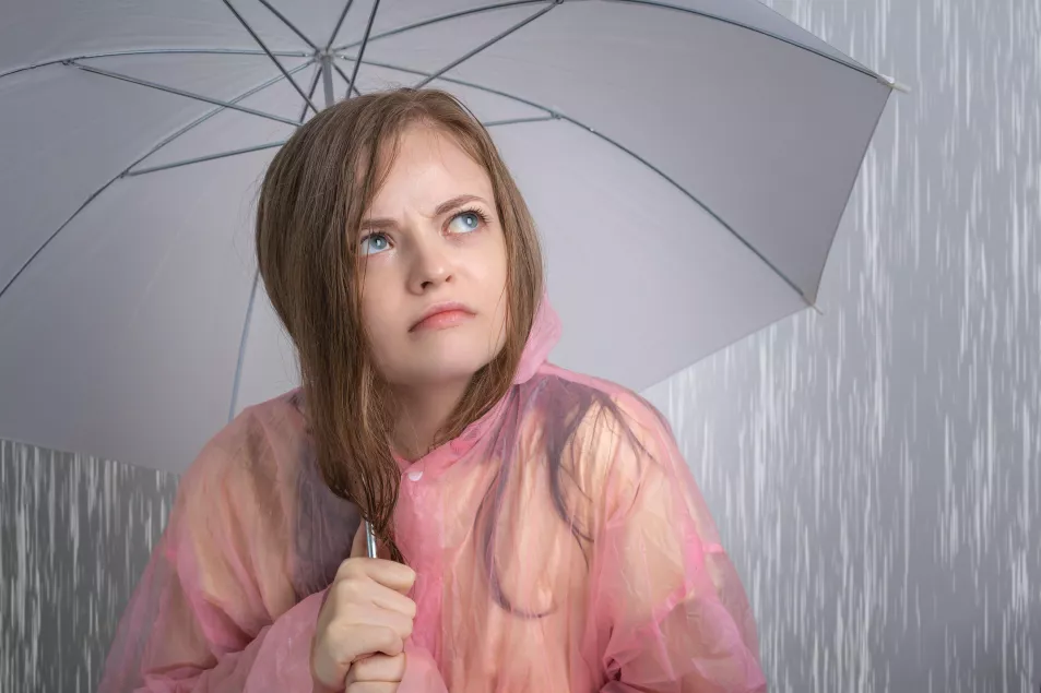 Young woman wearing a pink poncho looking unhappy under an umbrella while it rains 