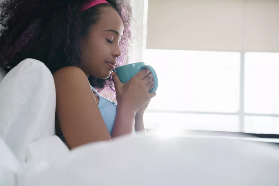 Woman sat up in bed smiling while sipping on a cup of coffee