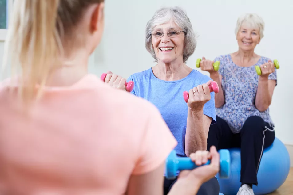 Two senior women sat on exercise balls lifting weights facing an instructor at a fitness class