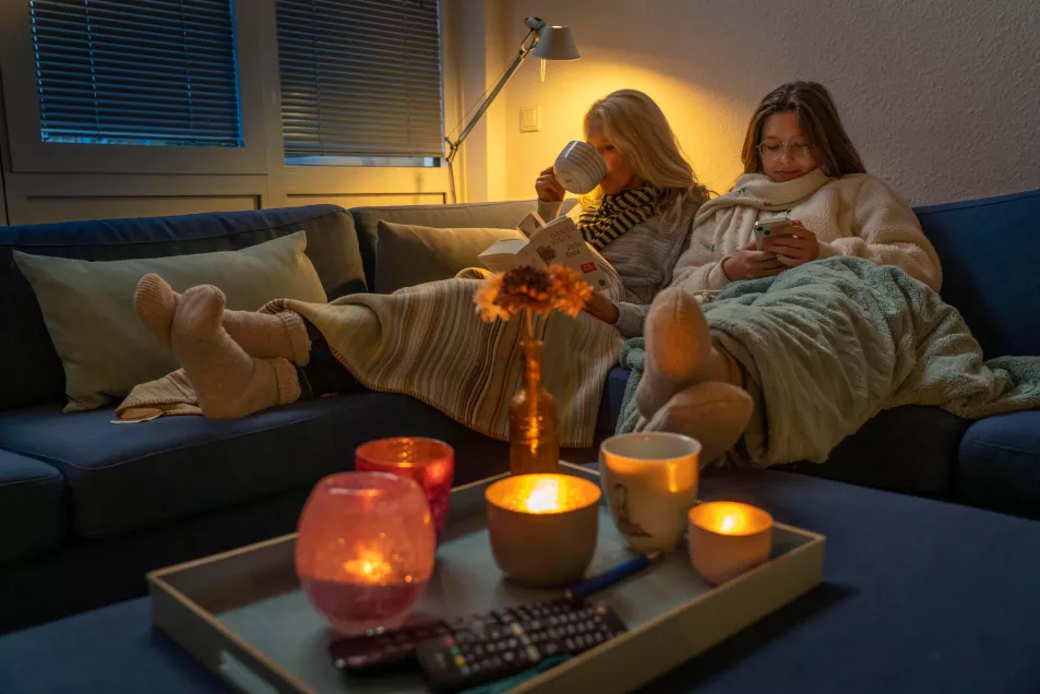 Two women sat in a candle lit living room looking cosy on a sofa while sipping hot drinks 