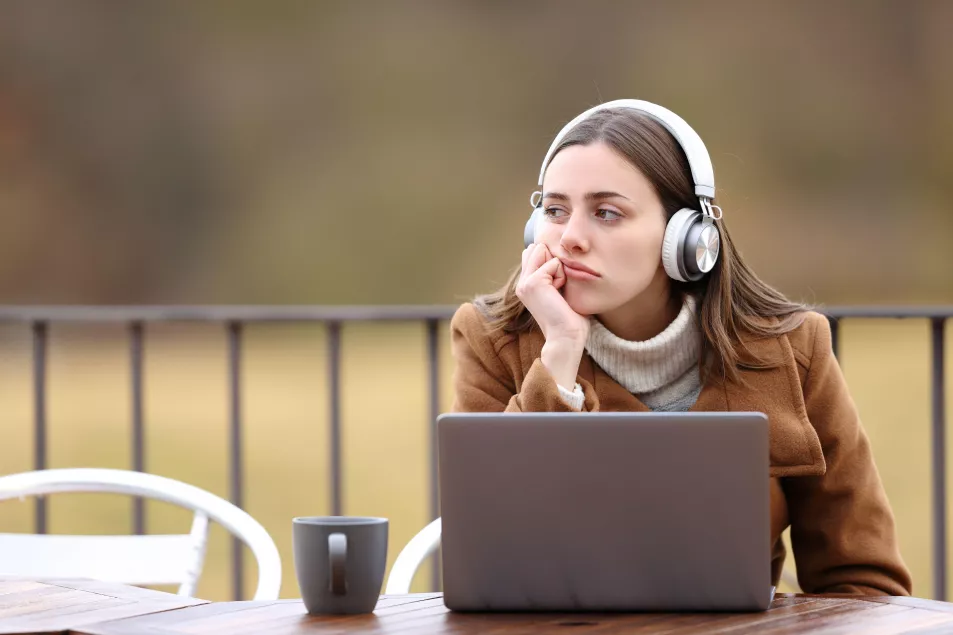 Woman wearing headphone and a coat sat outside a coffee shop looking bored with her laptop out