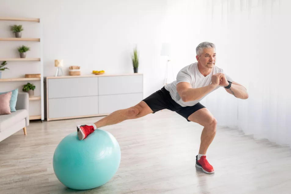 Athletic mature man doing side lunges, using fitness ball, working out at home in living room interior