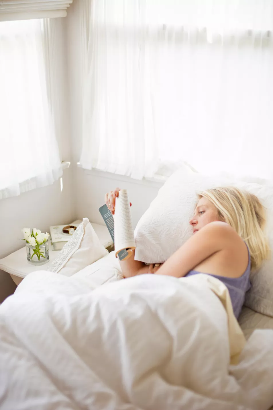 Blonde woman reading in a bed with white linen.