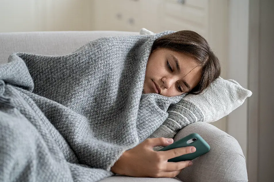 Moody sad teenage girl wrapped in a grey blanket lying on a sofa