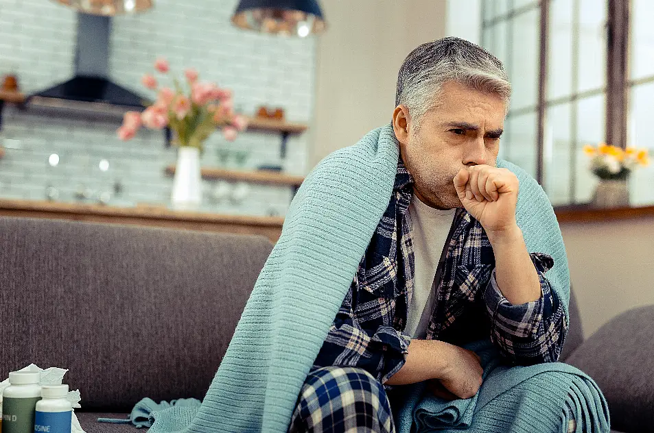 Middle aged man sat on a sofa coughing while wrapped in a blanket 