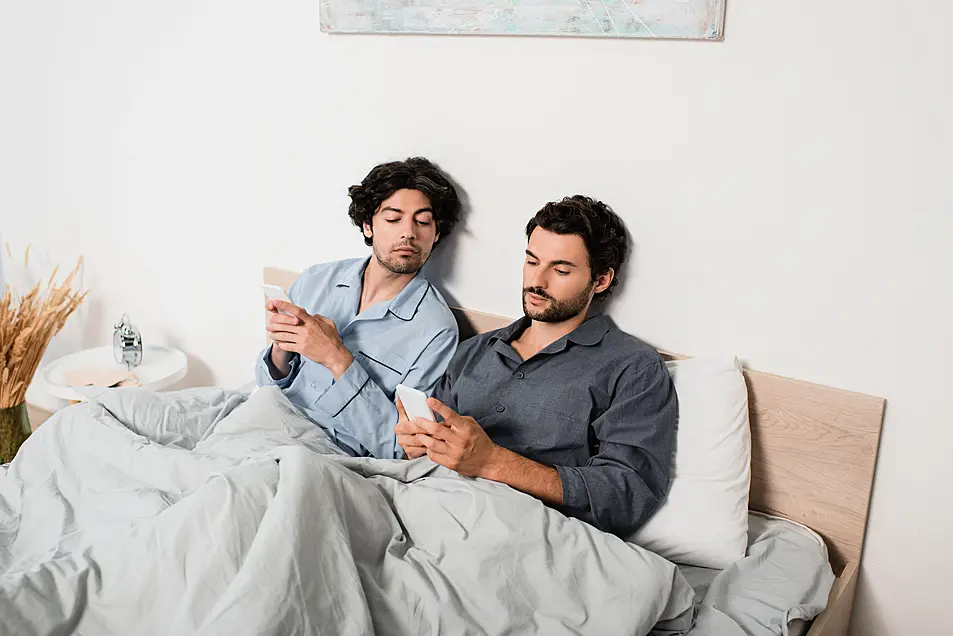 A gay male couple sitting in bed scrolling on their phones