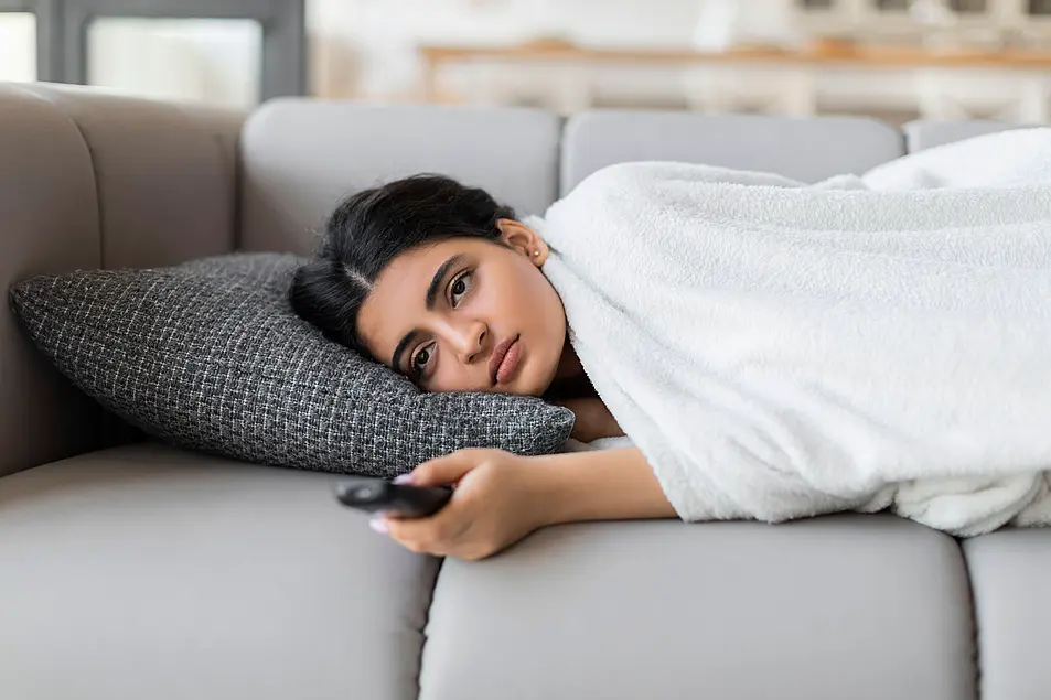 Woman laying on a grey sofa wrapped in a blanket pointing a remote at the TV