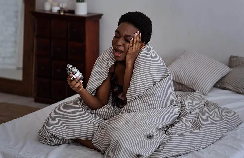 A woman sat up in bed yawning and looking at an alarm clock