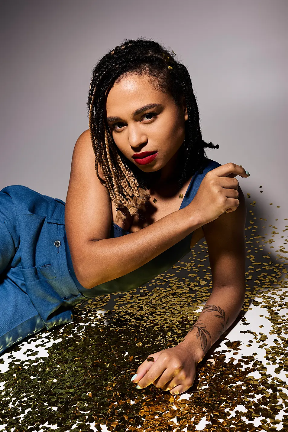 A chic young woman with braided hair and vibrant red lips showcases her unique style against a shimmering backdrop.