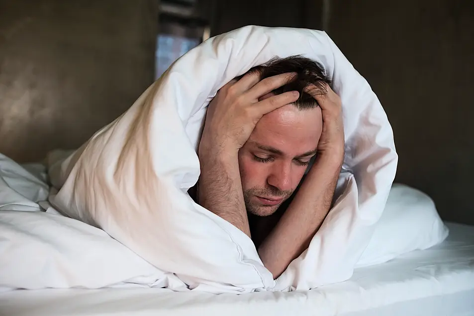 Depressed man lying in his bed wrapped in a duvet with his hands on his head