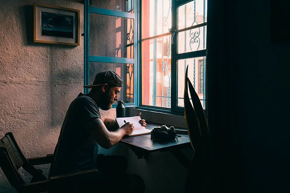 Male sits at wooden desk in a small dimly lit apartment 