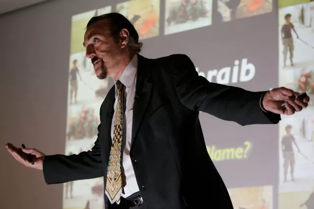 Dr Philip Zimbardo in a suit, with his arms outstretched in front of a projector