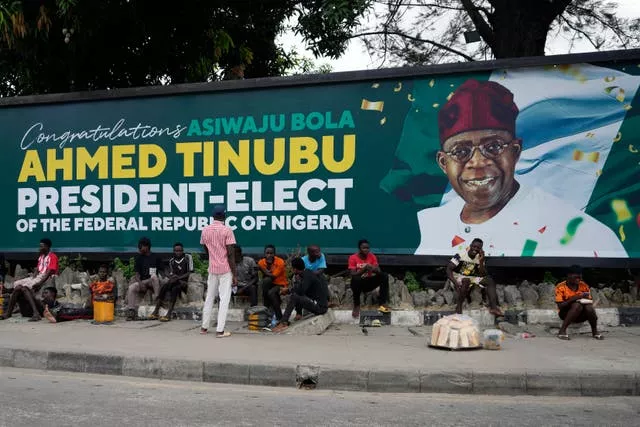 People sit under a billboard with a congratulatory message for the President-Elect Bola Ahmed Tinubu