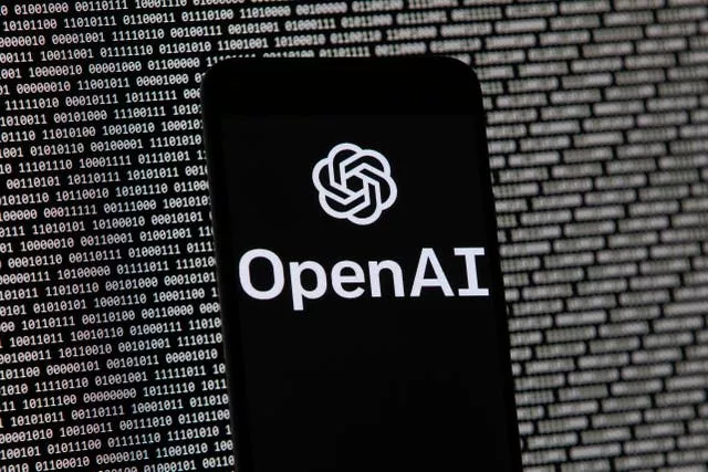 The OpenAI logo appears on a mobile phone in front of a computer screen