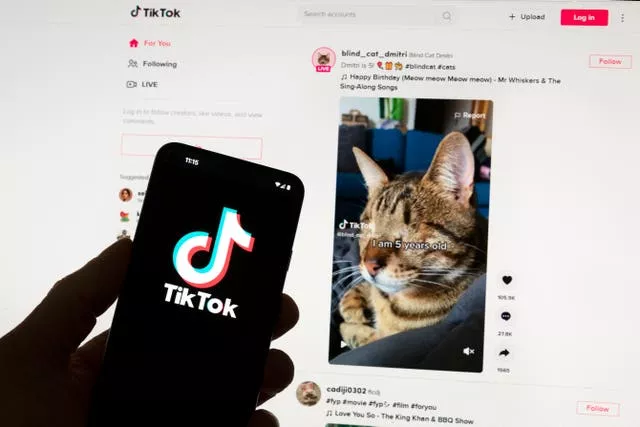 The TikTok logo seen on a mobile phone in front of a computer screen which displays the TikTok home screen 