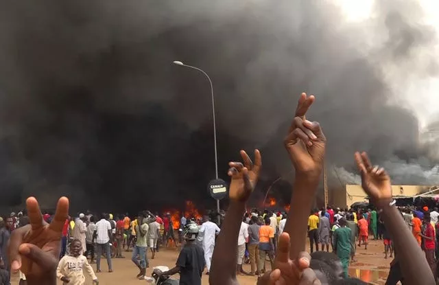 With the headquarters of the ruling party burning in the back, supporters of mutinous soldiers demonstrate in Niamey