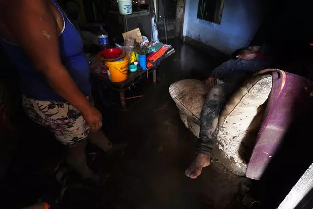 Elizabeth Morales shows her flood-damaged home as her husband lies on a waterlogged couch