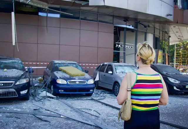 A blonde woman in a striped top and beige bag looks at damaged cars after Russia's missile attack in Dnipro. There is glass and metal covering the floor and red and white striped tape