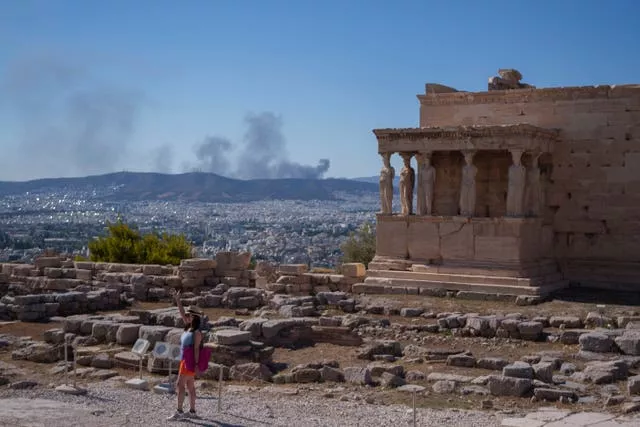 A woman poses for a photo in front Caryatid statues as smoke from a fire rises up in the background