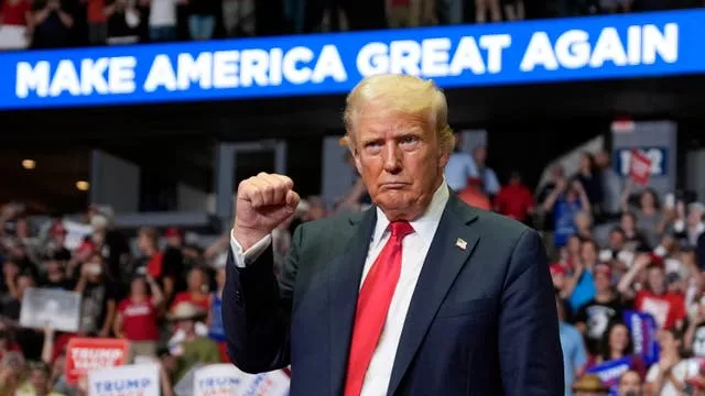 Donald Trump gestures at campaign rally with sign behind reading Make America Great Again