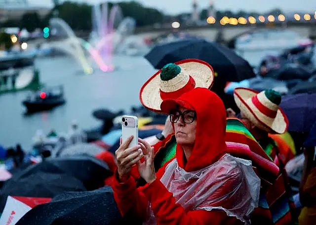 A spectator takes a photo on their phone as athletes ride in a boat along the Seine River during the opening ceremony for the Olympics in Paris