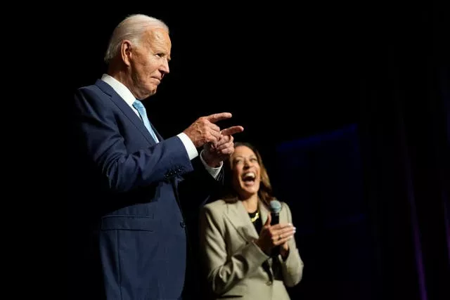 Joe Biden, in the foreground, points while Kamala Harris, in the background, laughs while holding a microphone