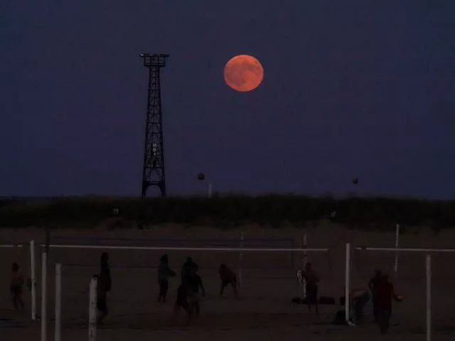 The pumpkin-coloured Harvest moon rises over the beach as people play volleyball on the shore of Lake Michigan