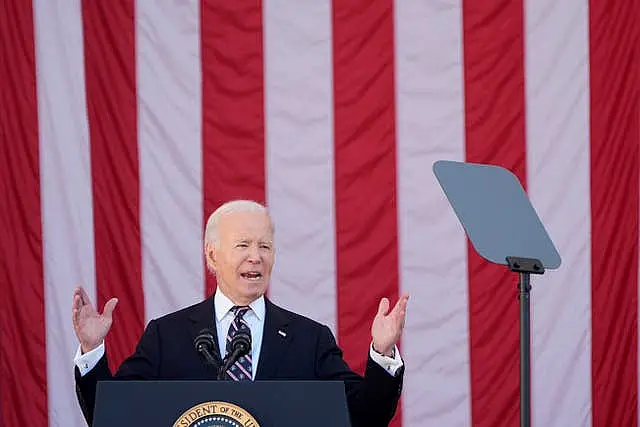 President Joe Biden raises his hands while speaking at Arlington National Cemetery, with an American flag in the background