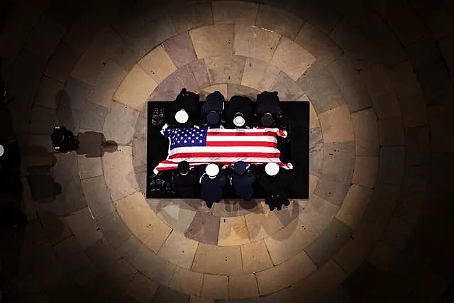 A bird's eye view looks down on the coffin draped in the American flag. A circle of light surrounds it, people are seated all around