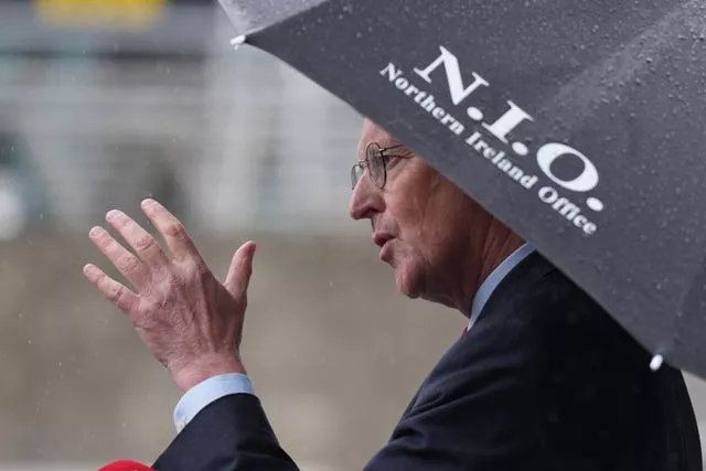 Northern Ireland Secretary Hilary Benn gestures with his hand while holding an umbrella saying Northern Ireland Office on it