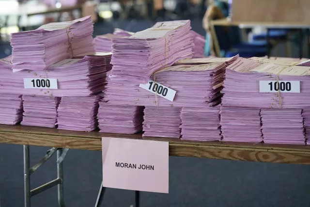 Stacks of pink ballots are grouped together with elastic bands in stacks of 1,000. There is a slip of paper tied to the table that says John Moran