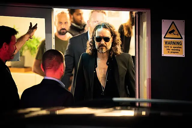 Russell Brand wearing dark glasses walking through a doorway to a waiting car.