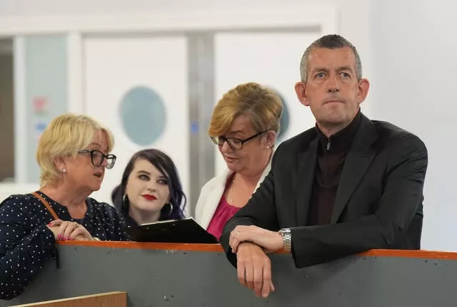 A man in a black suit and shirt stands with three women. The women are looking at a clipboard whilst the man is looking at someone out of shot