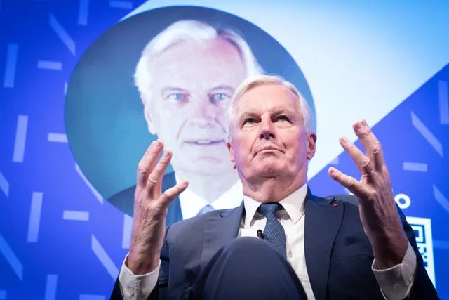 Michel Barnier sitting on stage with his image as a backdrop