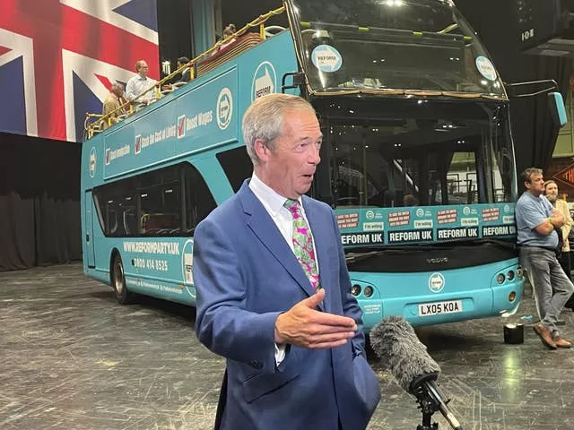 Nigel Farage standing in front of the Reform UK battlebus and speaking to a broadcast microphone