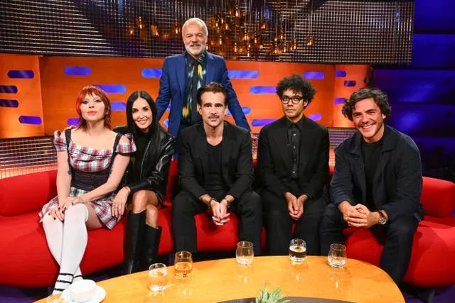 Graham Norton stands behind two women and three men who are sitting on a sofa