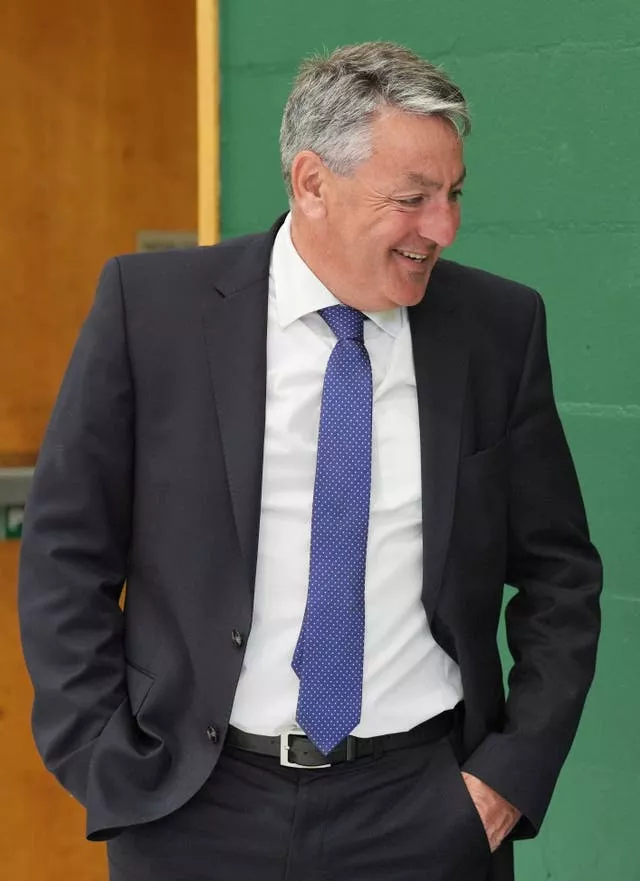 Man in black suit with blue tie is laughing at something out of shot. He has his hands in his pocket and his suit jacket unbuttoned