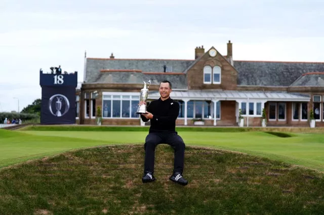 Xander Schauffele holding the Claret Jug, with his legs dangling over a bunker