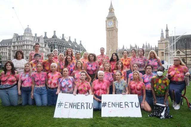 A group of women with painted breasts outside the Palace of Westminster holding signs reading hashtag Enhertu